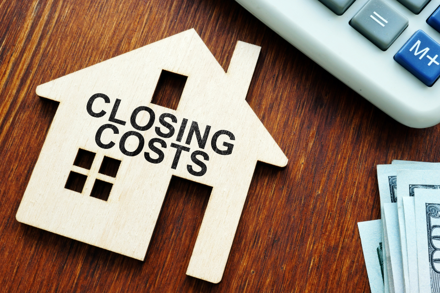 a wood cutout of a house with closing costs on front laying on a wooden desk with a calculator and money partially show in the corner